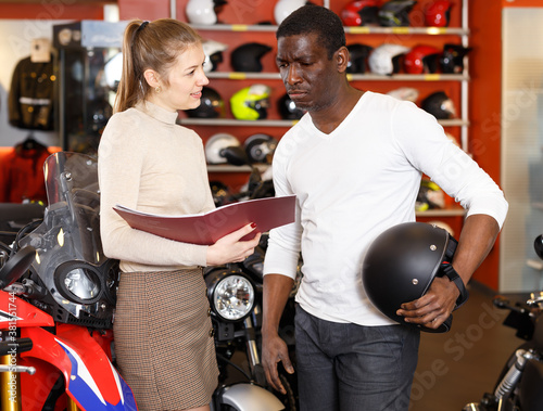 Smiling shopping assistant helping and demonstrating male customer motorcycles in store