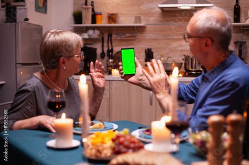 Elderly man and woman waving at phone with green screen. Aged people looking at mockup template chroma key isolated smart phone display using techology internet sitting at the table in kitchen.