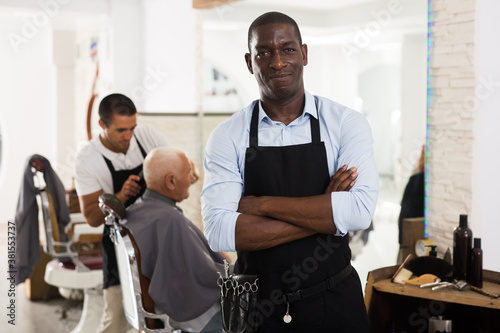 Confident African-American man hairdresser posing with folded arms in modern barber shop..