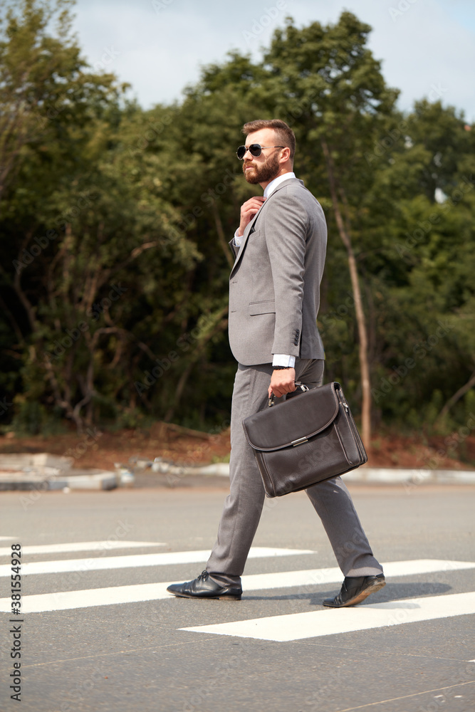 side view on caucasian businessman in suit or tuxedo walking with bag in hands, wearing sunglasses. in city streets side view on caucasian businessman in suit or tuxedo walking with bag in hands, wearing sunglasses. in city streets