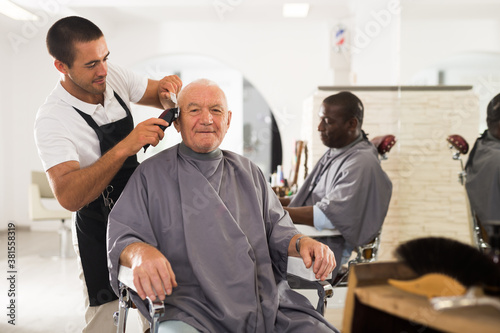 Elderly man getting haircut with electric clipper from skillful young barber in salon..