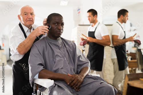 African-American man getting haircut with clipper from professional skillful elderly barber in hair salon
