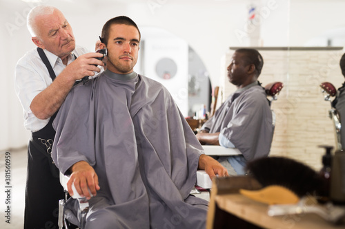 Young man getting haircut with clipper from professional skillful aged barber in salon