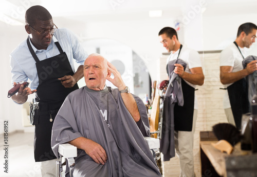 Dissatisfied elderly male client complaining about haircut to perplexed African hairdresser at barber shop