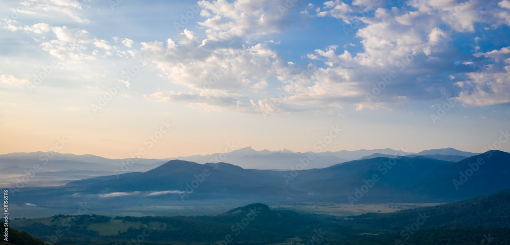 Early morning in the mountains. Morning sun and haze in the valley against the background of mountains. Mountain summer landscape. Banner size Early morning in the mountains. Morning sun and haze in the valley against the background of mountains. Mountain summer landscape. Banner size