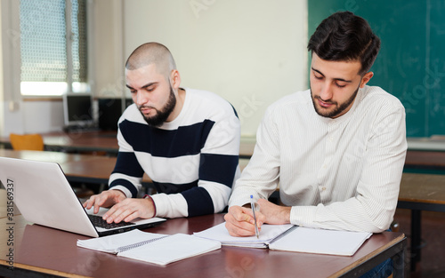 Two young handsome students working with laptop at desk in classroom