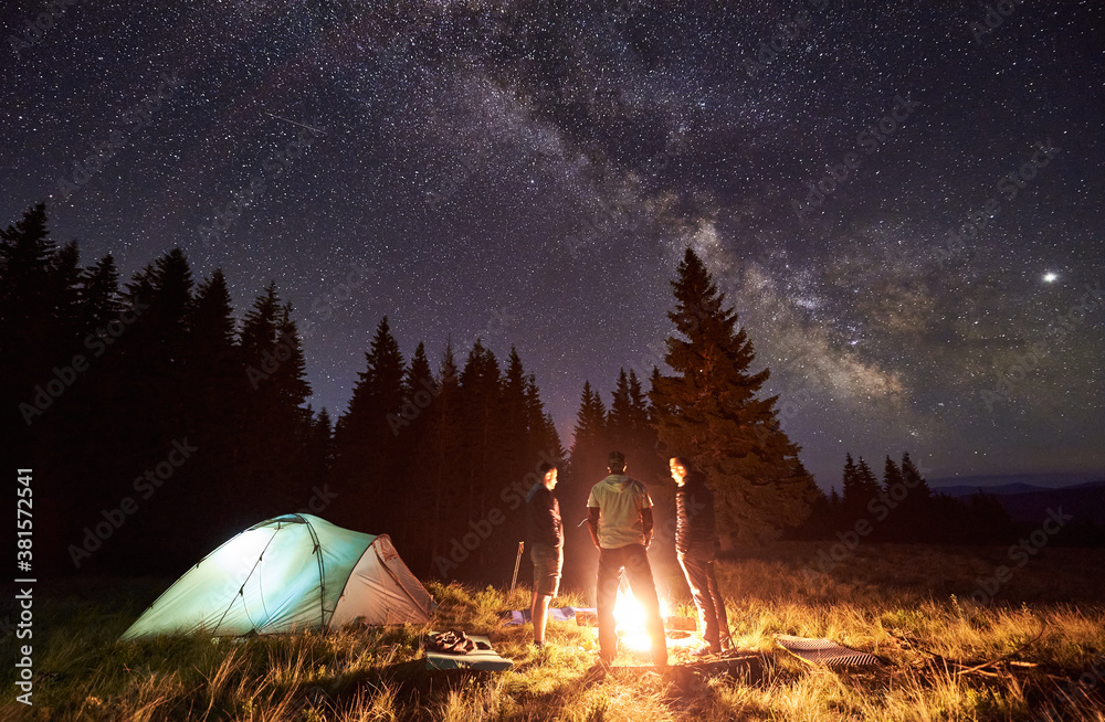Night summer camping in forest. Bright campfire burning, three male tourists standing around fire near tent under beautiful dark starry sky and Milky way. Concept of tourism, camping in the mountains. - obrazy, fototapety, plakaty 