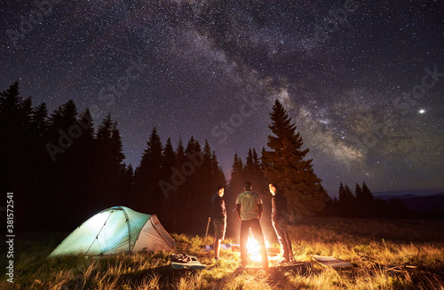 Night summer camping in forest. Bright campfire burning, three male tourists standing around fire near tent under beautiful dark starry sky and Milky way. Concept of tourism, camping in the mountains.
