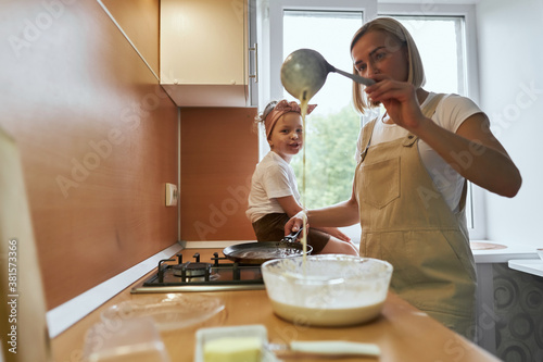 Fototapeta Naklejka Na Ścianę i Meble -  happy mother baking with little daughter in apron and cook hat working with flour , bowl and spoon preparing dough teaching the kid baking and having fun together.