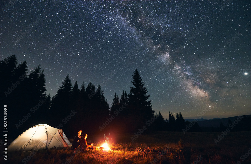Side view of loving couple sitting near bright burning campfire and tent, enjoying beautiful camping night together under dark sky full of shiny stars and bright Milky Way, warm summer night. - obrazy, fototapety, plakaty 