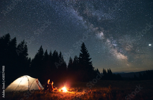 Side view of loving couple sitting near bright burning campfire and tent, enjoying beautiful camping night together under dark sky full of shiny stars and bright Milky Way, warm summer night.