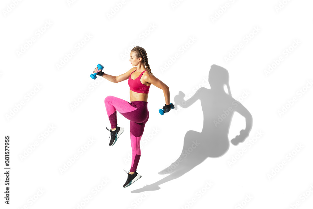 Weights. Beautiful young female athlete practicing on white studio background, portrait with shadows. Sportive fit model in motion and action. Body building, healthy lifestyle, style concept.