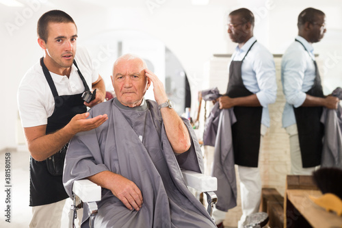 Portrait of shocked elderly man sitting in barber chair with confused young hairdresser behind him