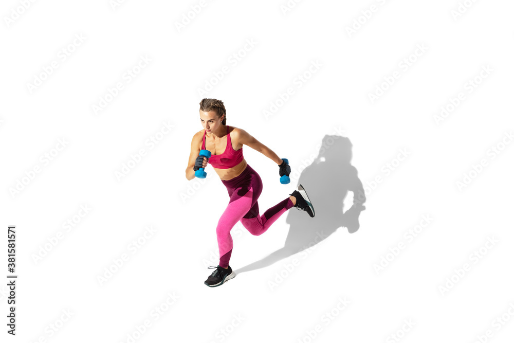 Running. Beautiful young female athlete practicing on white studio background, portrait with shadows. Sportive fit model in motion and action. Body building, healthy lifestyle, style concept.