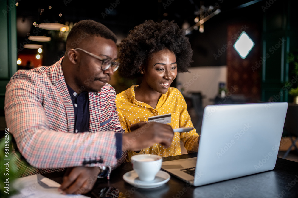 Attractive smiling african couple sitting in cafe and purchasing over internet things for house.