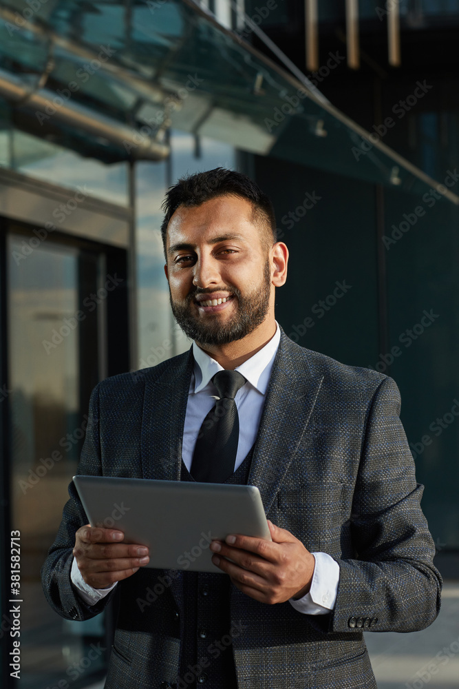 Portrait of bearded businessman in elegant suit smiling at camera while using tablet pc outdoors