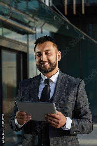Portrait of bearded businessman in elegant suit smiling at camera while using tablet pc outdoors