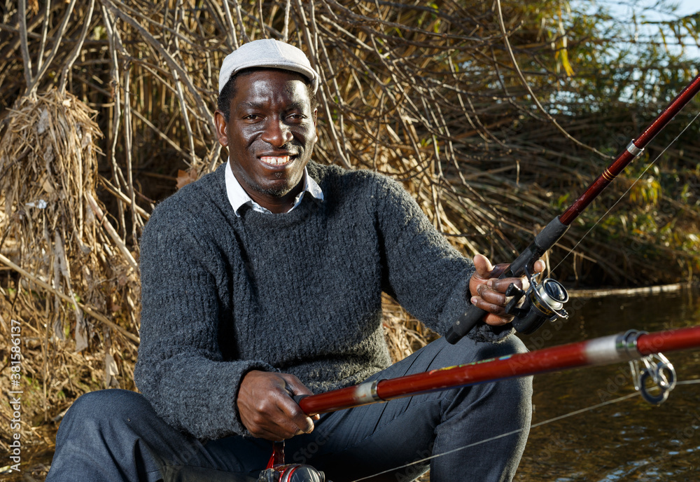 Fototapeta premium Portrait of cheerful afro fisherman sitting on wooden stool and fishing