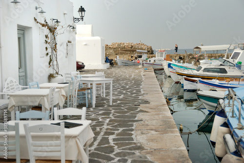 Puste stoliki restauracji w porcie rybackim na greckiej wyspie Paros. Łodzi rybackie przycumowane na wprost stolików.