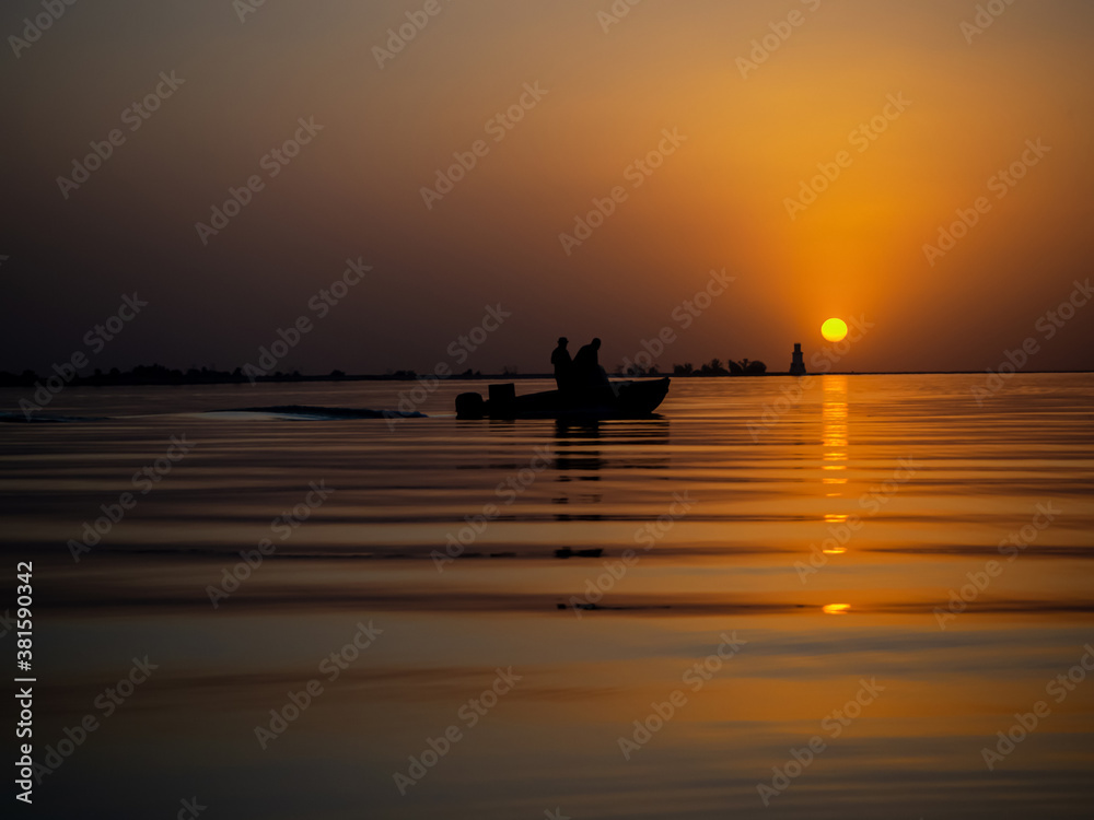 Fishermen in a fishing boat on background of a Golden sunset on sea. Full calm, calm surface of water in ocean with a view of the lighthouse. Reflection of the orange dawn sun on waves. Copy space