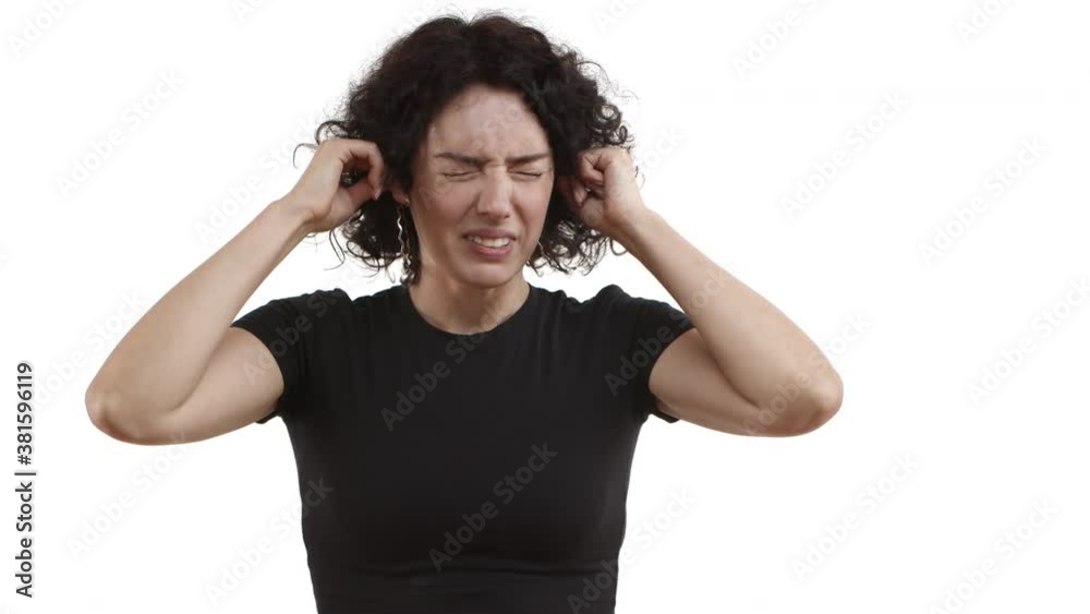 Young woman with curly short hair, wearing black t-shirt, plug fingers in ears, shaking head in denial, grimacing bothered by loud noise, standing over white background
