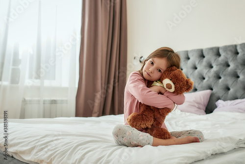A little girl embracing a teddybear while sitting cross-legged barefoot on a huge bed