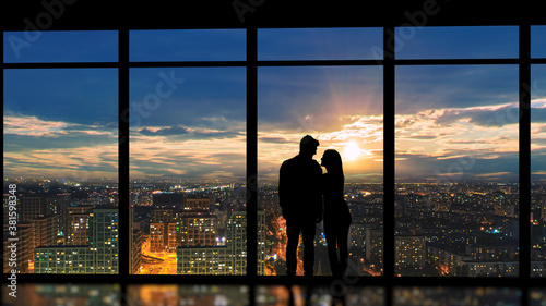 The couple standing near the panoramic window against the beautiful sunset