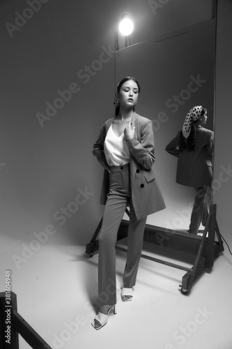 Elegant fashionable woman in brown suit, sabot shoes and polka dot kerchief standing and posing in studio on gray background between mirrors. Fashion portrait with reflections. Beautiful teenager girl