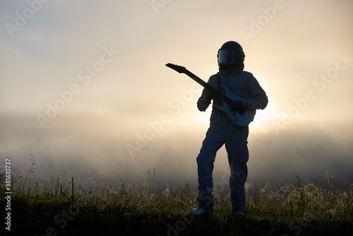 Silhouette of space traveler playing melody on guitar in misty grassy valley with white mystical sky on background. Cosmonaut guitarist with musical instrument wearing while space suit and helmet.