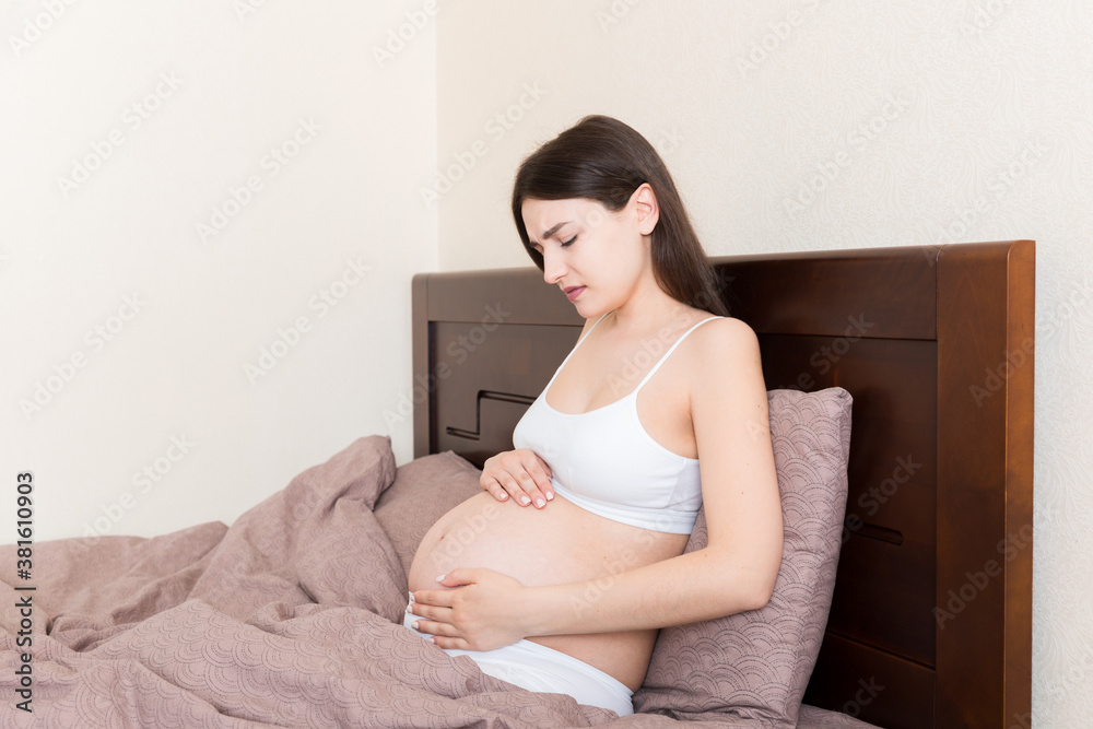 Young pretty pregnant woman, in pain, sitting on bed in bedroom, preparing to give a birth Young pretty pregnant woman, in pain, sitting on bed in bedroom, preparing to give a birth