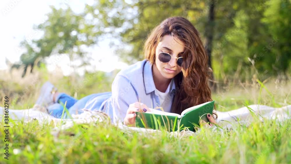 Beautiful young lady with curly dark hair lying on soft blanket and reading interesting book at summer park. Cute female relaxing on fresh air with new literature. Beautiful young lady with curly dark hair lying on soft blanket and reading interesting book at summer park. Cute female relaxing on fresh air with new literature.