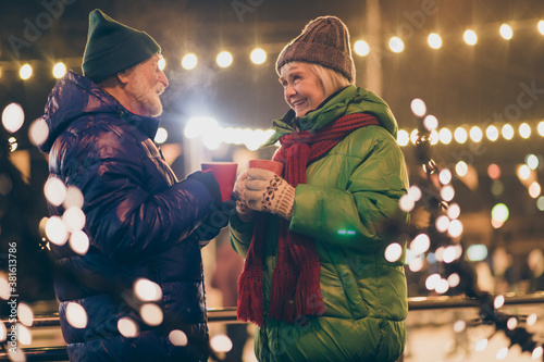 Photo of two people pensioner couple hold mugs drink eggnog comfortable cheerful smile newyear atmosphere wear mittens coat red scarf headwear x-mas night street park lights fair outside