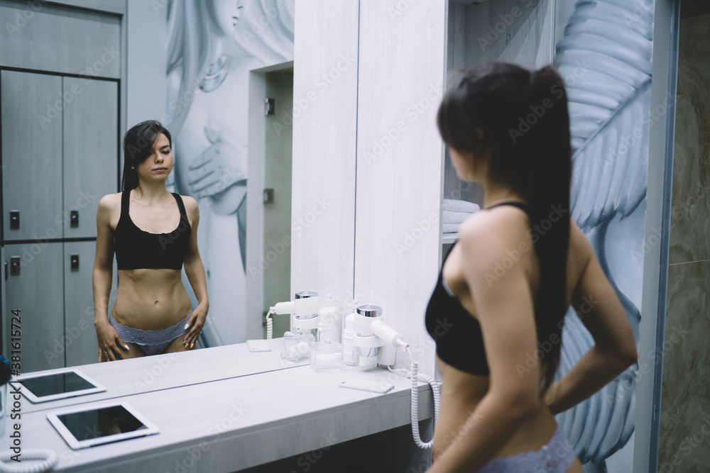 Back view of female with slim figure in lingerie looking at mirror satisfied with slimming results, beautiful caucasian young woman enjoying training prepare for workout in modern locker room Back view of female with slim figure in lingerie looking at mirror satisfied with slimming results, beautiful caucasian young woman enjoying training prepare for workout in modern locker room