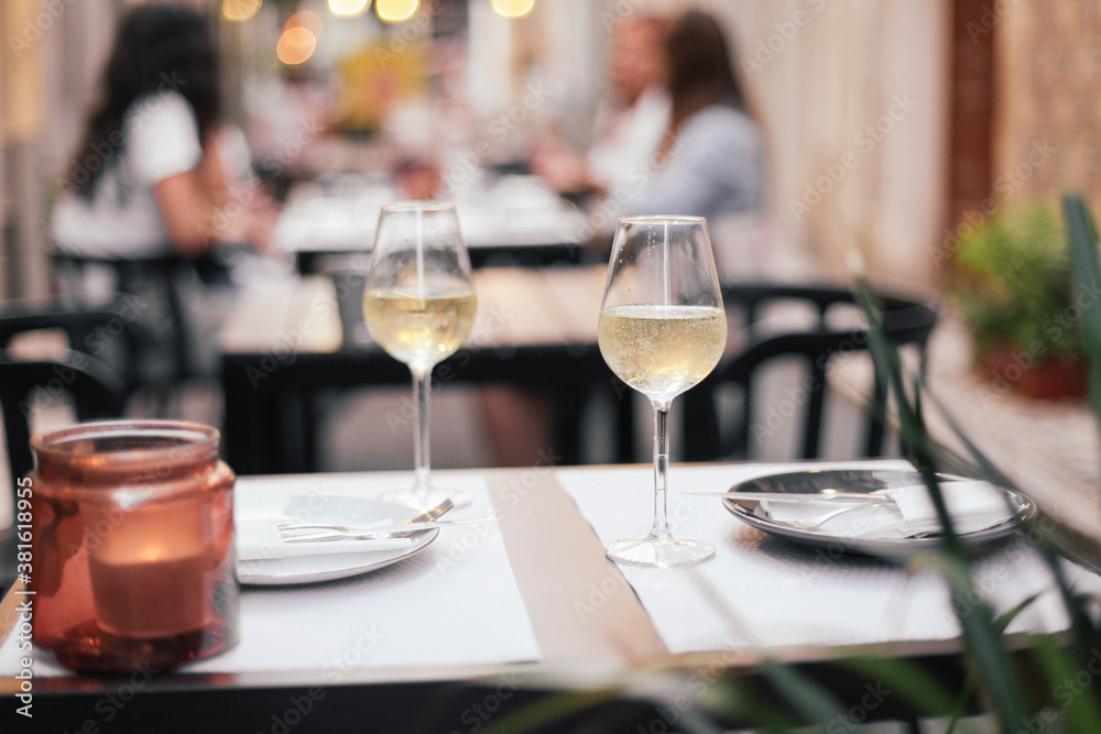 Two glasses of white wine on a table in an outdoor terrace, romantic dinner date with evening cozy lights Two glasses of white wine on a table in an outdoor terrace, romantic dinner date with evening cozy lights