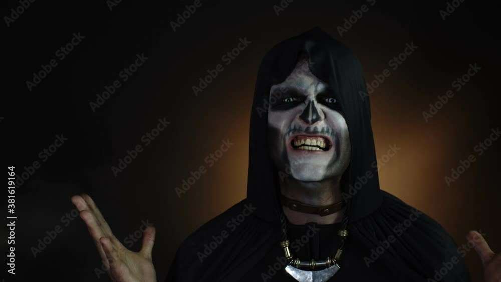 Sinister man with skeleton makeup in hood making angry faces, looking at camera, trying to scare Sinister man with skeleton makeup in hood making angry faces, looking at camera, trying to scare