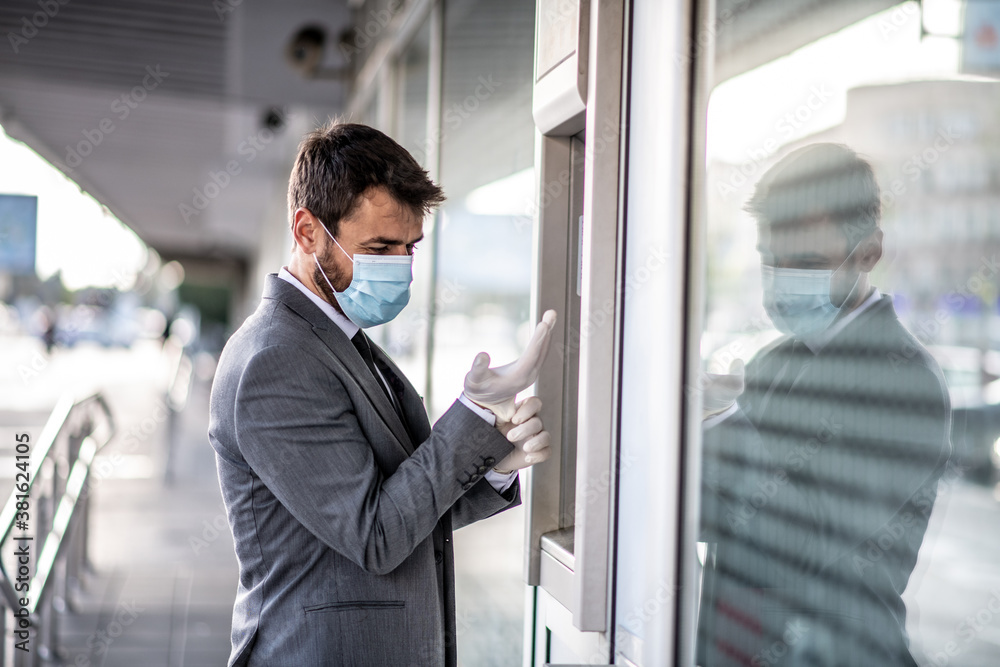 A young businessman with a mask puts on another glove, stands in front of cash machine, life during a pandemic caused by a virus A young businessman with a mask puts on another glove, stands in front of cash machine, life during a pandemic caused by a virus