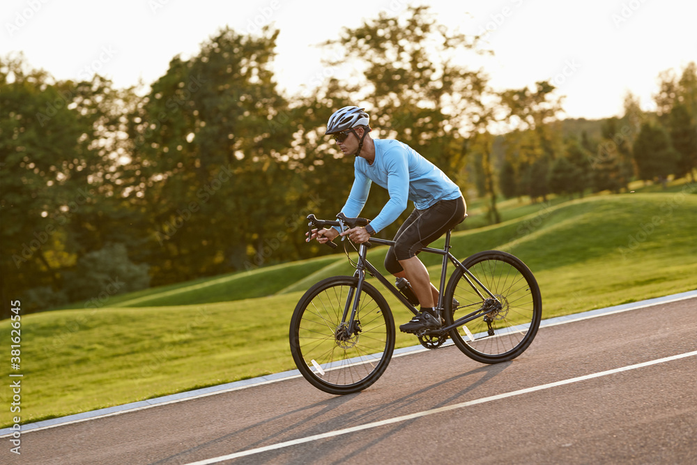 Obraz premium Side view of a young athletic man in sportswear, professional bicycle racer riding mountain road bike in the city park at sunset