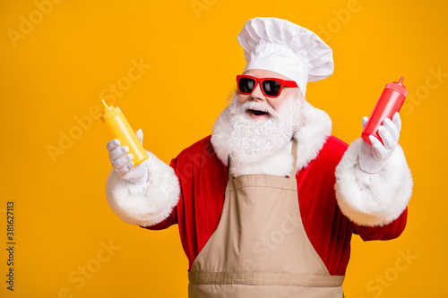 Photo of positive santa claus chef hold mustard tomato ketchup sauce bottle x-mas eve noel snack wear headwear sunglass apron isolated over yellow shine color background