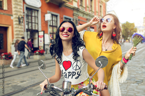 two young beautiful hipster women riding on motorbike city street, summer europe vacation, traveling, smiling, happy, having fun, sunglasses, stylish outfit, adventures, positive, friends together