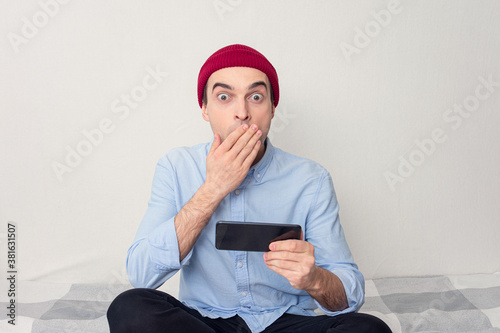 Surprised guy is sitting at home, young man is reading a text message on his phone, portrait photo