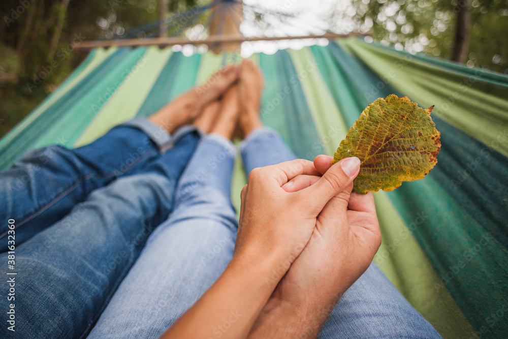 Obraz premium man and woman in love lying in hammock embracing, legs, close-up details, romance, autumn vacation, forest, barefoot, holding leaf in hands together, jeans, denim outfit