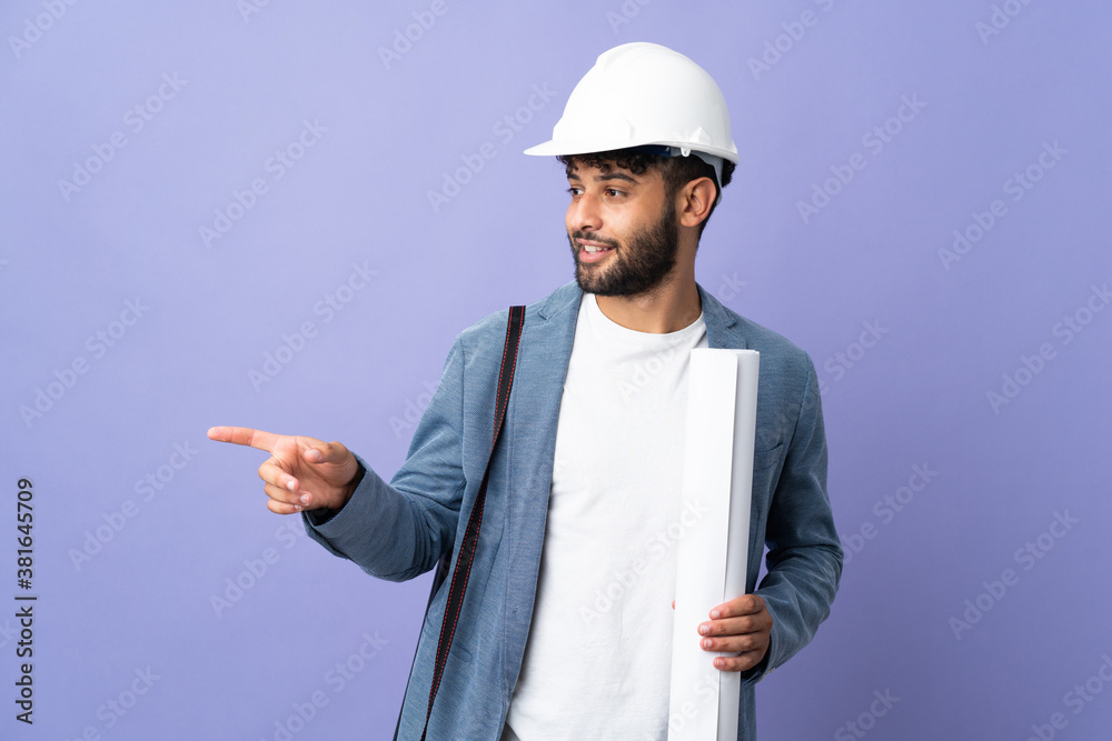 Young architect Moroccan man with helmet and holding blueprints over isolated background pointing finger to the side Young architect Moroccan man with helmet and holding blueprints over isolated background pointing finger to the side