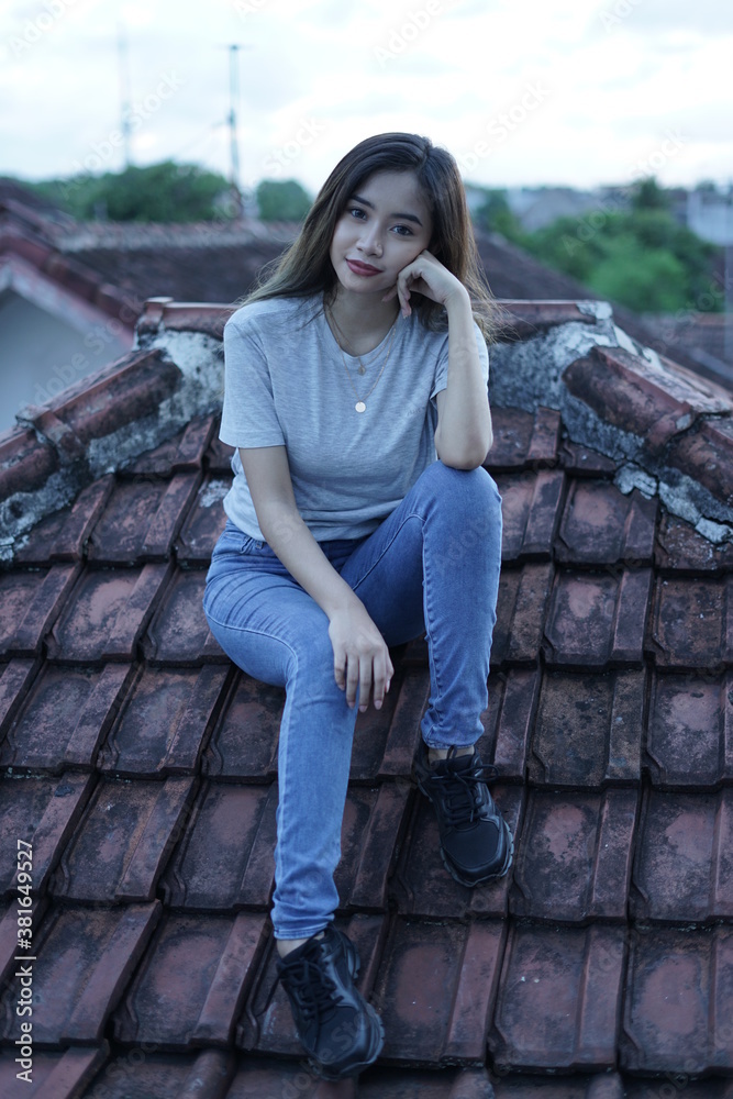 The blonde girl sits on the roof tile while stylishly wearing a gray shirt. female t-shirt models for mockups and templates. The blonde girl sits on the roof tile while stylishly wearing a gray shirt. female t-shirt models for mockups and templates.