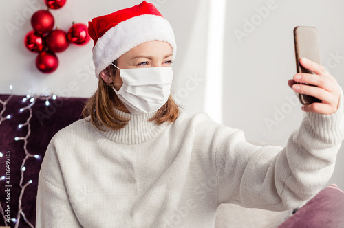 Young woman in a white sweater, a Santa Claus hat and a medical mask talking by video link with relatives and friends