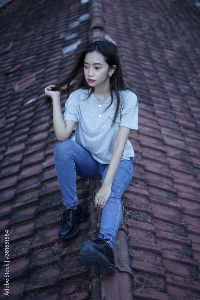 The blonde girl sits on the roof tile while stylishly wearing a gray shirt. female t-shirt models for mockups and templates.