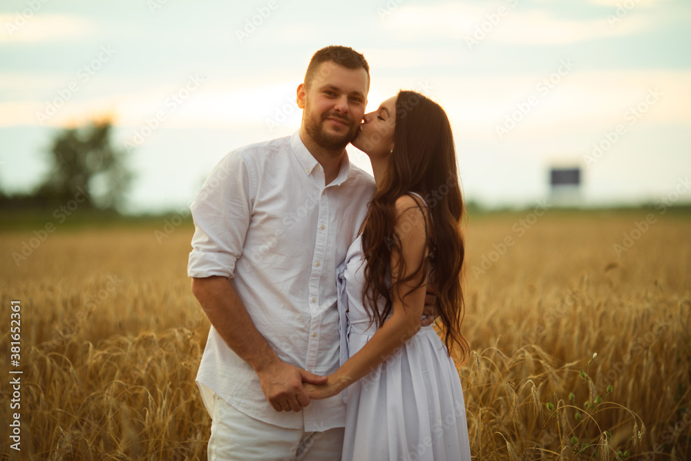 Smiling man hugging her pretty wife while standing behind her on a wheat field during evening sunset. Love concept