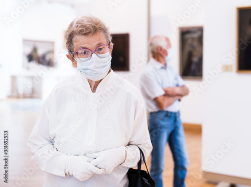 elderly European woman in mask protecting against covid examines paintings on display in hall of art museum