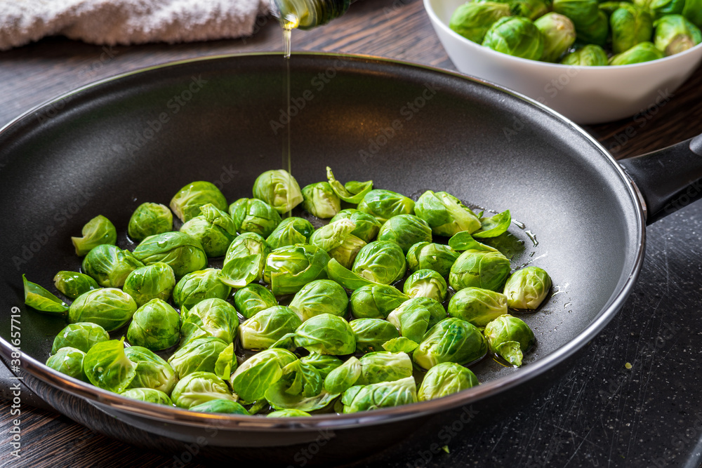 Fresh cut brussels sprouts in a pan with olive oil drizzle above them on a wooden table. White bowl and towel in the background.
