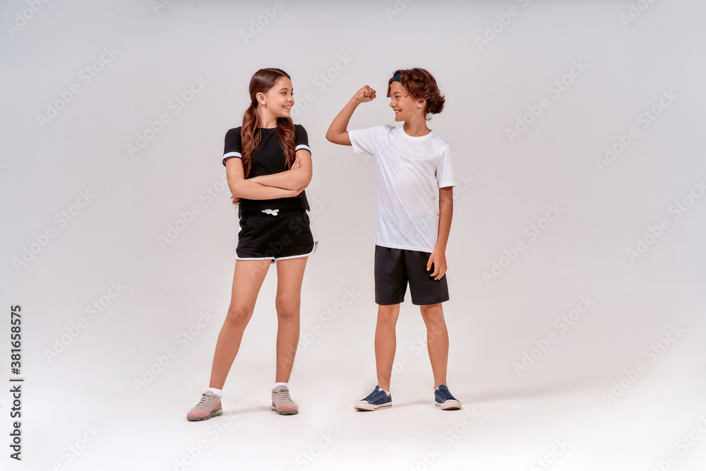 Two teenagers exercising together. Boy showing his biceps to a cute girl, they standing isolated over grey background in studio Two teenagers exercising together. Boy showing his biceps to a cute girl, they standing isolated over grey background in studio