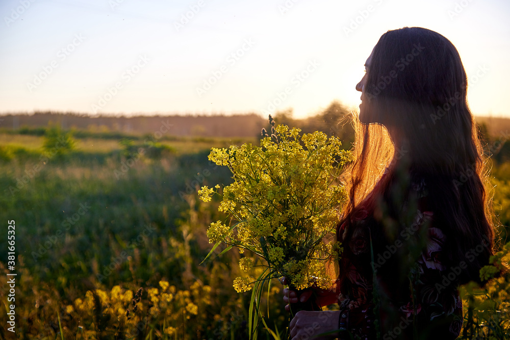 Fototapeta premium Beautiful woman in red dress holding bouquet of yellow flowers and enjoying summer sunny day in green field with green grass in sunset with nice orange sun